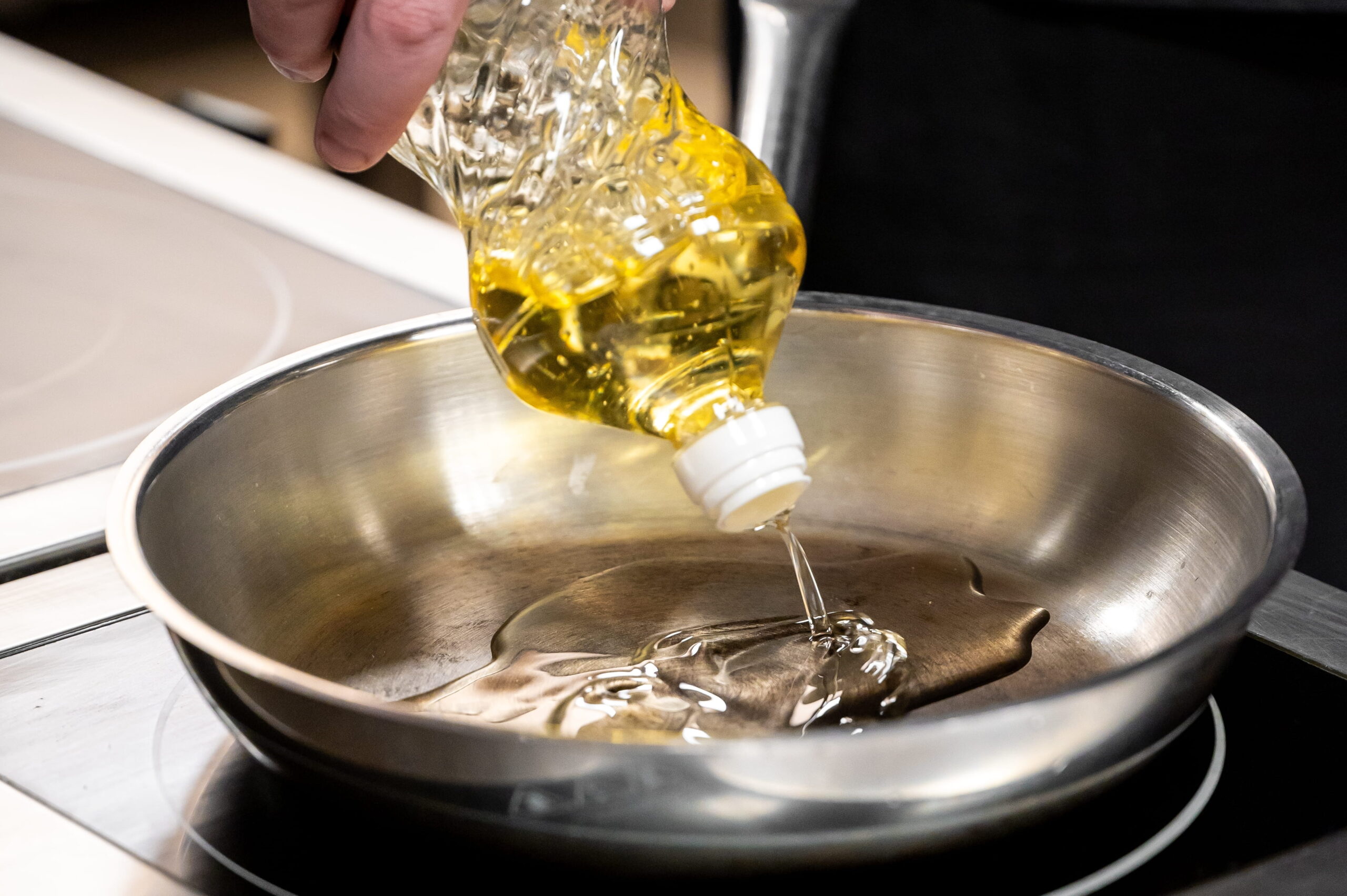 a hand pours golden cooking oil from a plastic bottle into a stainless steel frying pan